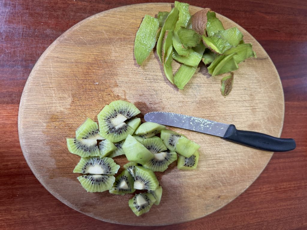 Chopping board with kiwi fruit peelings and sliced up fruit next to a black handled knife